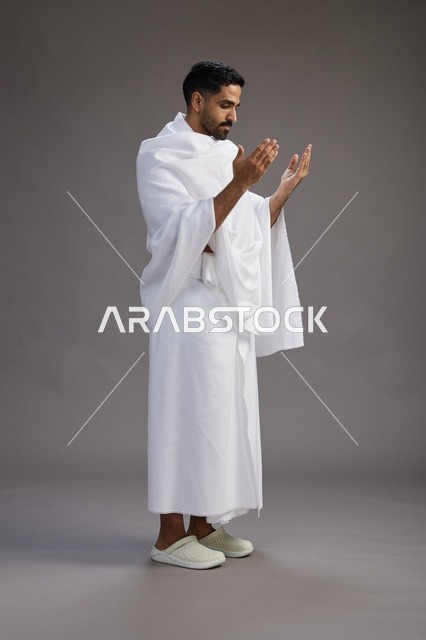 بورتريه لرجل سعودي بملابس الإحرام يدعو رافعاً يديه, الدعاء والتضرع لله , كامل الجسم, خلفيه رماديه