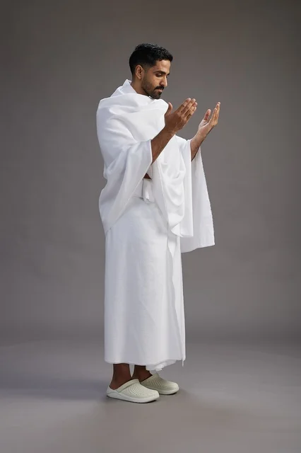 بورتريه لرجل سعودي بملابس الإحرام يدعو رافعاً يديه, الدعاء والتضرع لله , كامل الجسم, خلفيه رماديه