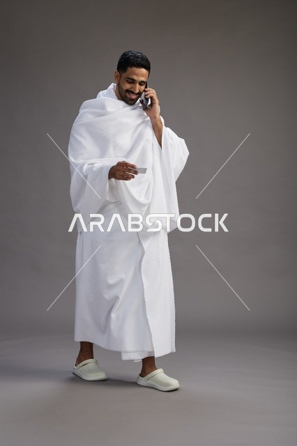 بورتريه لرجل سعودي بملابس الإحرام يحمل بطاقة بيضاء فارغه, مفهموم الحج والعمره, كامل الجسم, خلفيه رماديه