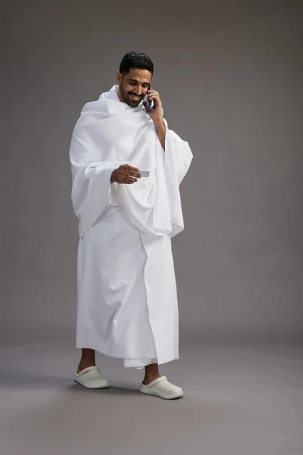 A portrait of a Saudi man in Ihram clothing holding a blank white card, representing the concepts of Hajj and Umrah, full body, gray background.