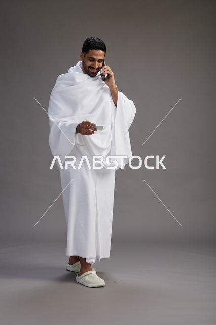 بورتريه لرجل سعودي بملابس الإحرام يحمل بطاقة بيضاء فارغه, مفهموم الحج والعمره, كامل الجسم, خلفيه رماديه