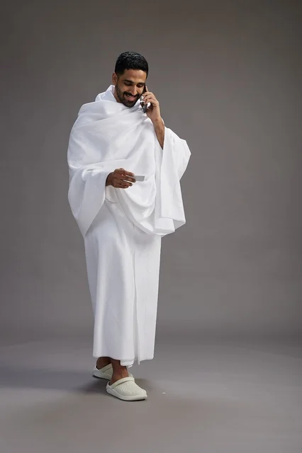 A portrait of a Saudi man in Ihram clothing holding a blank white card, representing the concepts of Hajj and Umrah, full body, gray background.