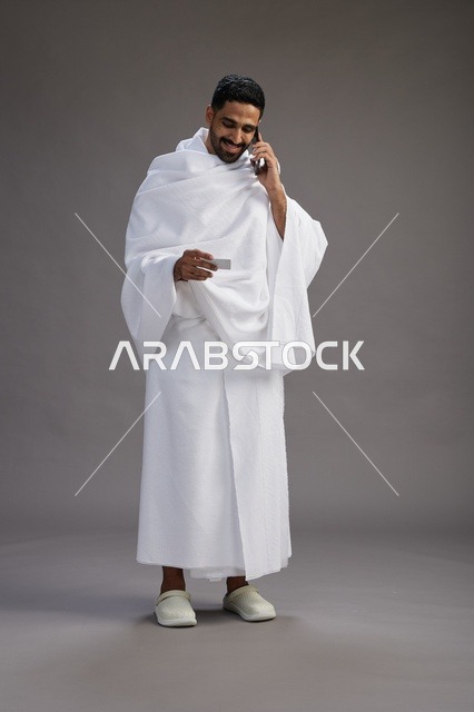 بورتريه لرجل سعودي بملابس الإحرام يحمل بطاقة بيضاء فارغه, مفهموم الحج والعمره, كامل الجسم, خلفيه رماديه