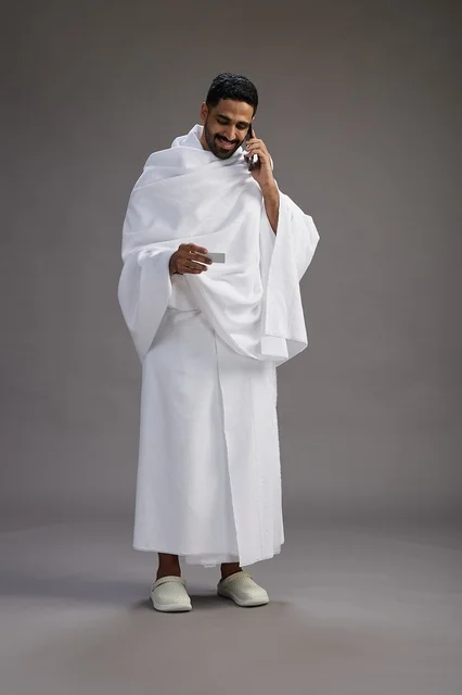 بورتريه لرجل سعودي بملابس الإحرام يحمل بطاقة بيضاء فارغه, مفهموم الحج والعمره, كامل الجسم, خلفيه رماديه