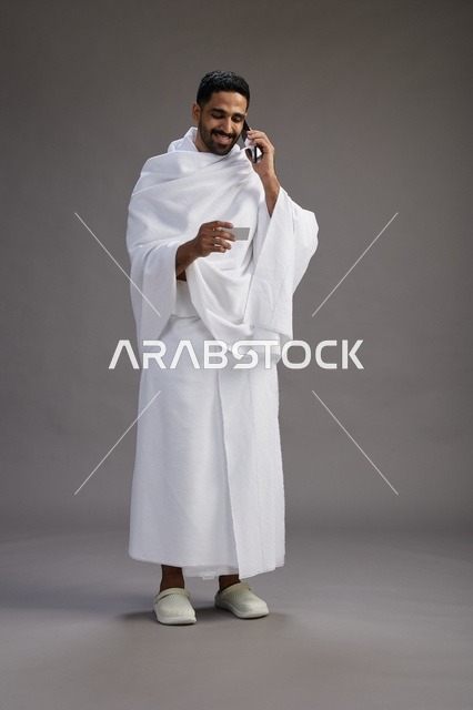 بورتريه لرجل سعودي بملابس الإحرام يحمل بطاقة بيضاء فارغه, مفهموم الحج والعمره, كامل الجسم, خلفيه رماديه