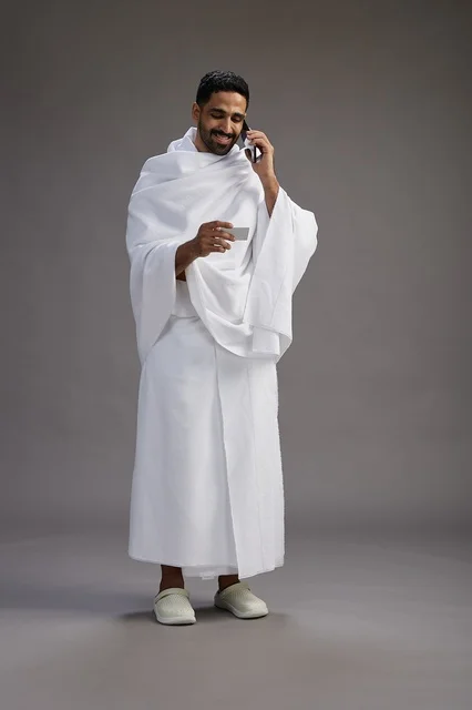 A portrait of a Saudi man in Ihram clothing holding a blank white card, representing the concepts of Hajj and Umrah, full body, gray background. A portrait of a Saudi man in Ihram clothing holding a blank white card, representing the concepts of Hajj and Umrah, full body, gray background.