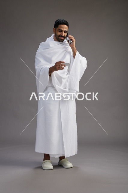 بورتريه لرجل سعودي بملابس الإحرام يحمل بطاقة بيضاء فارغه, مفهموم الحج والعمره, كامل الجسم, خلفيه رماديه