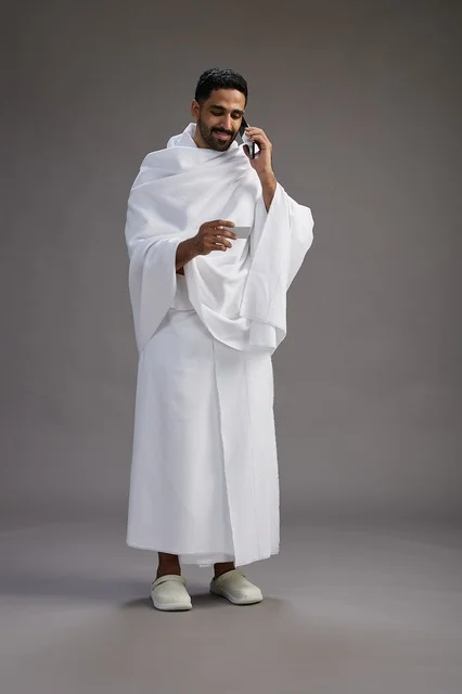 A portrait of a Saudi man in Ihram clothing holding a blank white card, representing the concepts of Hajj and Umrah, full body, gray background.