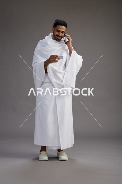 بورتريه لرجل سعودي بملابس الإحرام يحمل بطاقة بيضاء فارغه, مفهموم الحج والعمره, كامل الجسم, خلفيه رماديه