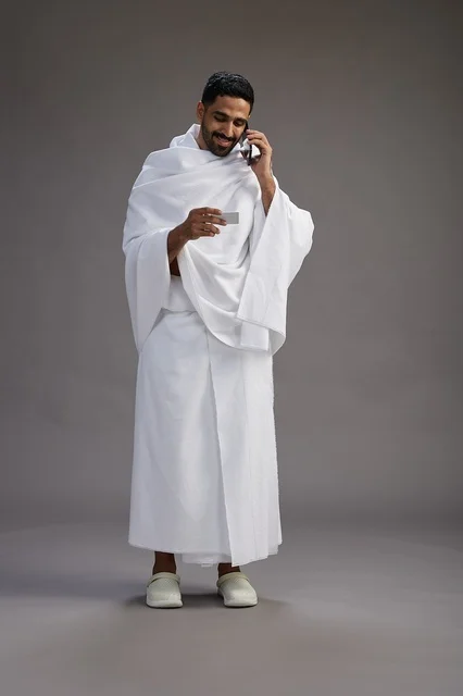 A portrait of a Saudi man in Ihram clothing holding a blank white card, representing the concepts of Hajj and Umrah, full body, gray background. A portrait of a Saudi man in Ihram clothing holding a blank white card, representing the concepts of Hajj and Umrah, full body, gray background.