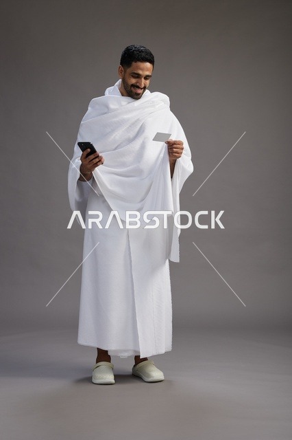بورتريه لرجل سعودي بملابس الإحرام يحمل بطاقة بيضاء فارغه, مفهموم الحج والعمره, كامل الجسم, خلفيه رماديه