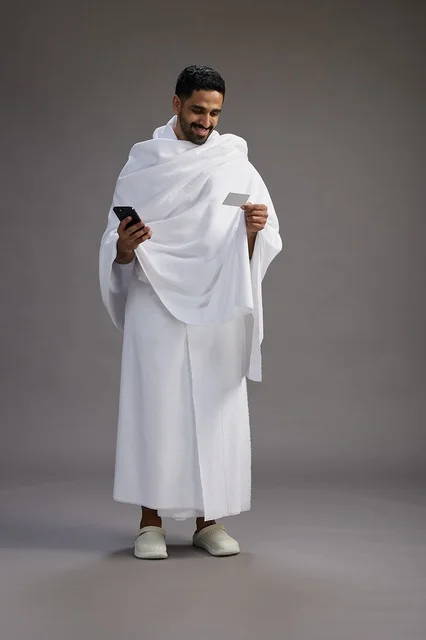 A portrait of a Saudi man in Ihram clothing holding a blank white card, representing the concepts of Hajj and Umrah, full body, gray background.