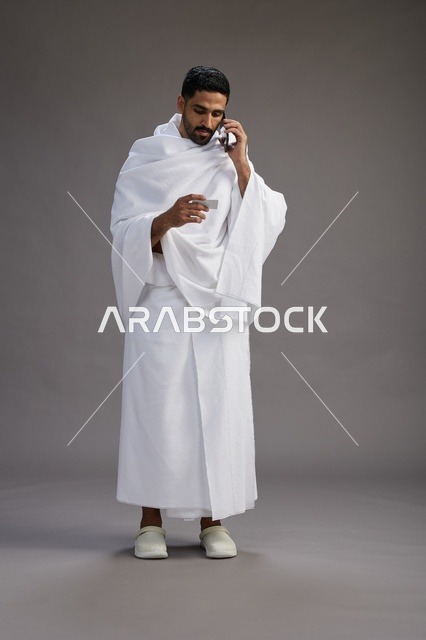 A portrait of a Saudi man in Ihram clothing holding a blank white card, representing the concepts of Hajj and Umrah, full body, gray background.