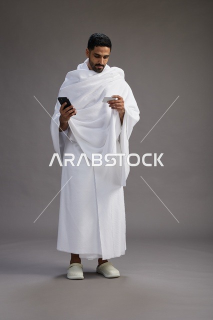 بورتريه لرجل سعودي بملابس الإحرام يحمل بطاقة بيضاء فارغه, مفهموم الحج والعمره, كامل الجسم, خلفيه رماديه
