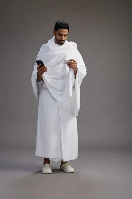 A portrait of a Saudi man in Ihram clothing holding a blank white card, representing the concepts of Hajj and Umrah, full body, gray background.
