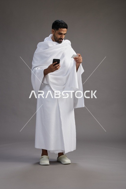 بورتريه لرجل سعودي بملابس الإحرام يحمل بطاقة بيضاء فارغه, مفهموم الحج والعمره, كامل الجسم, خلفيه رماديه
