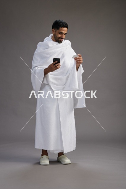 بورتريه لرجل سعودي بملابس الإحرام يحمل بطاقة بيضاء فارغه, مفهموم الحج والعمره, كامل الجسم, خلفيه رماديه