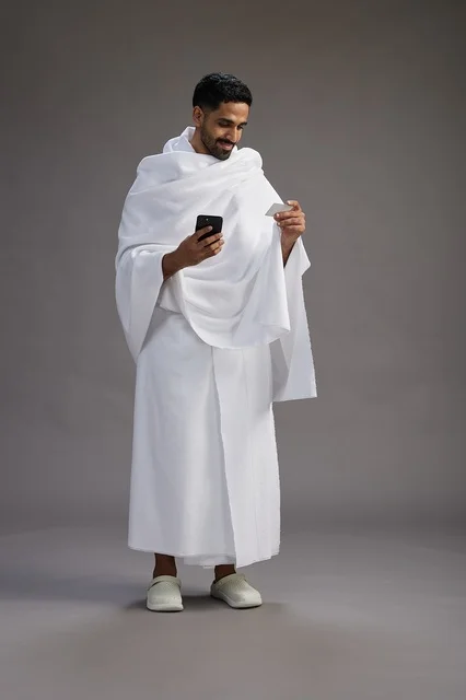 A portrait of a Saudi man in Ihram clothing holding a blank white card, representing the concepts of Hajj and Umrah, full body, gray background.