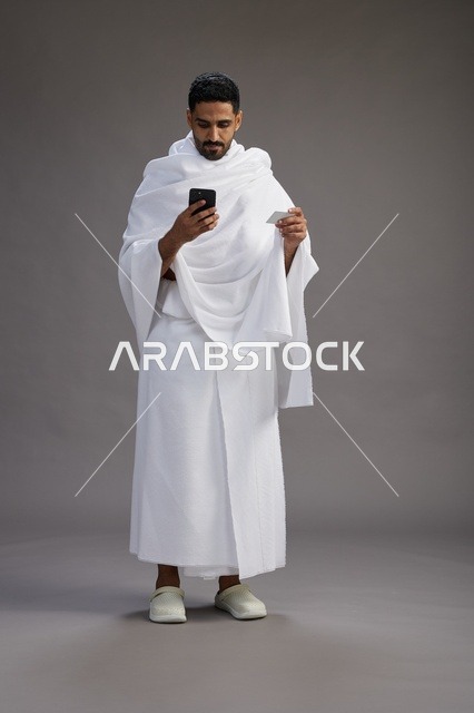 بورتريه لرجل سعودي بملابس الإحرام يحمل بطاقة بيضاء فارغه, مفهموم الحج والعمره, كامل الجسم, خلفيه رماديه