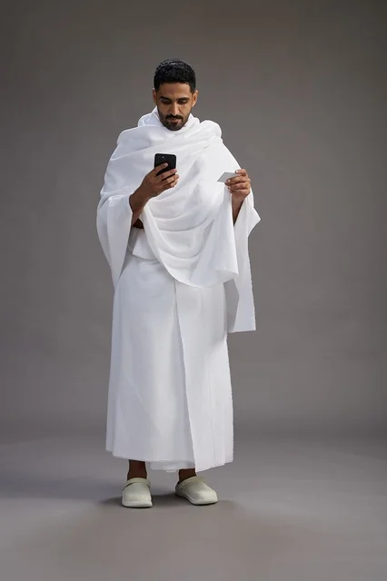بورتريه لرجل سعودي بملابس الإحرام يحمل بطاقة بيضاء فارغه, مفهموم الحج والعمره, كامل الجسم, خلفيه رماديه