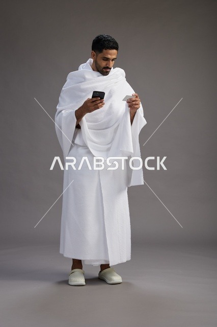 بورتريه لرجل سعودي بملابس الإحرام يحمل بطاقة بيضاء فارغه, مفهموم الحج والعمره, كامل الجسم, خلفيه رماديه