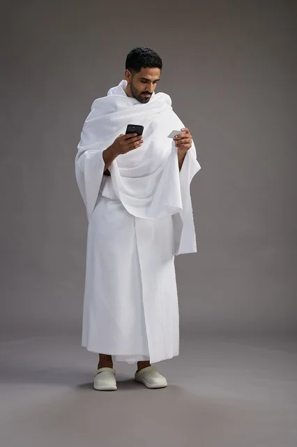 بورتريه لرجل سعودي بملابس الإحرام يحمل بطاقة بيضاء فارغه, مفهموم الحج والعمره, كامل الجسم, خلفيه رماديه