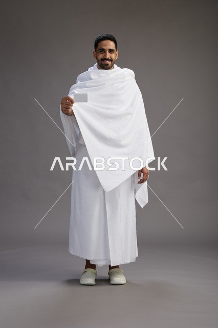 A portrait of a Saudi man in Ihram clothing holding a blank white card, representing the concepts of Hajj and Umrah, full body, gray background.