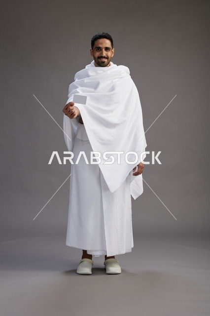 بورتريه لرجل سعودي بملابس الإحرام يحمل بطاقة بيضاء فارغه, مفهموم الحج والعمره, كامل الجسم, خلفيه رماديه