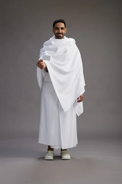 A portrait of a Saudi man in Ihram clothing holding a blank white card, representing the concepts of Hajj and Umrah, full body, gray background.