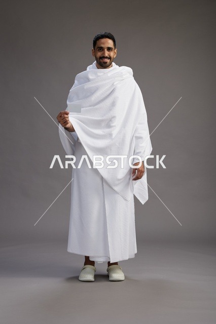 بورتريه لرجل سعودي بملابس الإحرام يحمل بطاقة بيضاء فارغه, مفهموم الحج والعمره, كامل الجسم, خلفيه رماديه