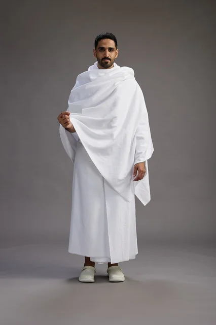 بورتريه لرجل سعودي بملابس الإحرام يحمل بطاقة بيضاء فارغه, مفهموم الحج والعمره, كامل الجسم, خلفيه رماديه