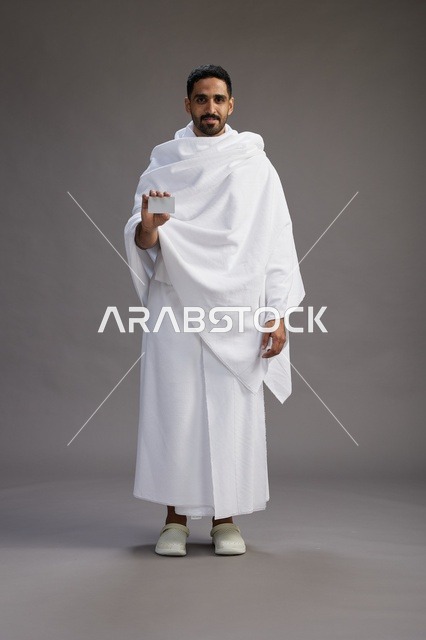 بورتريه لرجل سعودي بملابس الإحرام يحمل بطاقة بيضاء فارغه, مفهموم الحج والعمره, كامل الجسم, خلفيه رماديه