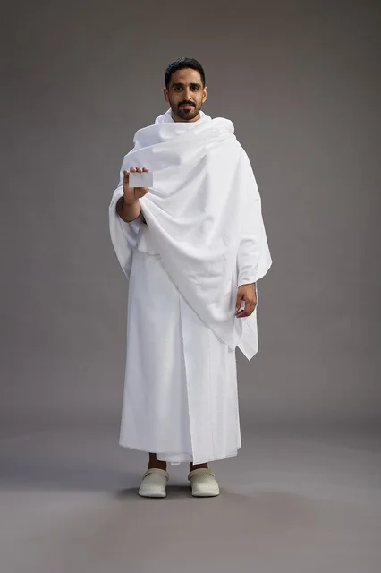 A portrait of a Saudi man in Ihram clothing holding a blank white card, representing the concepts of Hajj and Umrah, full body, gray background. A portrait of a Saudi man in Ihram clothing holding a blank white card, representing the concepts of Hajj and Umrah, full body, gray background.