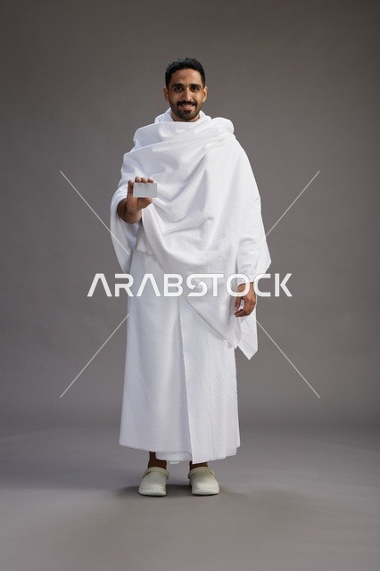 بورتريه لرجل سعودي بملابس الإحرام يحمل بطاقة بيضاء فارغه, مفهموم الحج والعمره, كامل الجسم, خلفيه رماديه