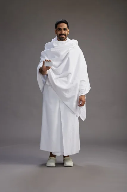 A portrait of a Saudi man in Ihram clothing holding a blank white card, representing the concepts of Hajj and Umrah, full body, gray background.