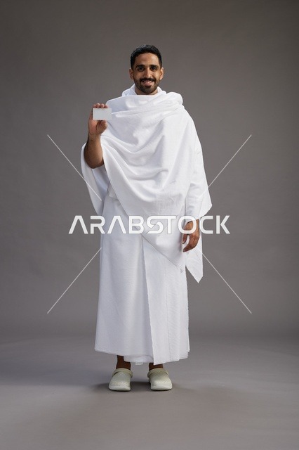 بورتريه لرجل سعودي بملابس الإحرام يحمل بطاقة بيضاء فارغه, مفهموم الحج والعمره, كامل الجسم, خلفيه رماديه