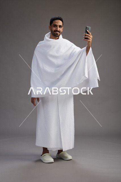 بورتريه لرجل سعودي بملابس الإحرام يبتسم ويلتقط صورة سيلفي, استخدام الهاتف المحمول, الحج والعمره في مكه, الجسم بالكامل, خلفيه رماديه