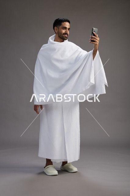 بورتريه لرجل سعودي بملابس الإحرام يبتسم ويلتقط صورة سيلفي, استخدام الهاتف المحمول, الحج والعمره في مكه, الجسم بالكامل, خلفيه رماديه