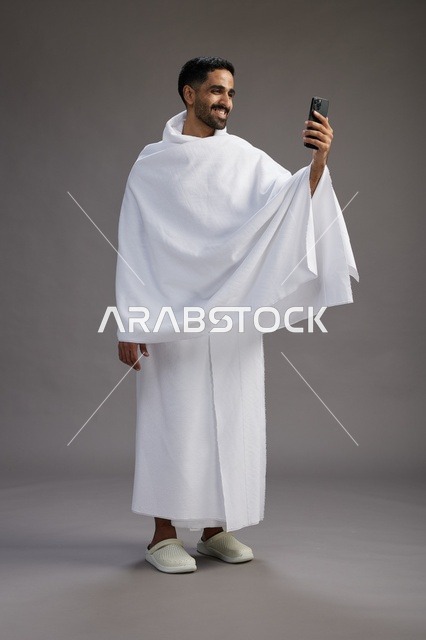 بورتريه لرجل سعودي بملابس الإحرام يبتسم ويلتقط صورة سيلفي, استخدام الهاتف المحمول, الحج والعمره في مكه, الجسم بالكامل, خلفيه رماديه