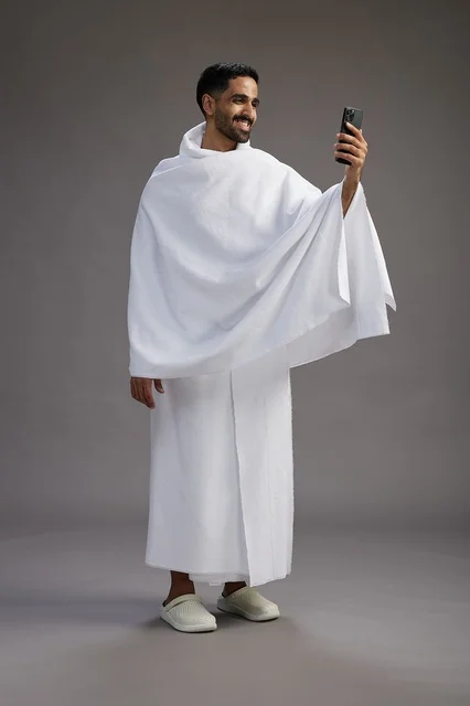 بورتريه لرجل سعودي بملابس الإحرام يبتسم ويلتقط صورة سيلفي, استخدام الهاتف المحمول, الحج والعمره في مكه, الجسم بالكامل, خلفيه رماديه