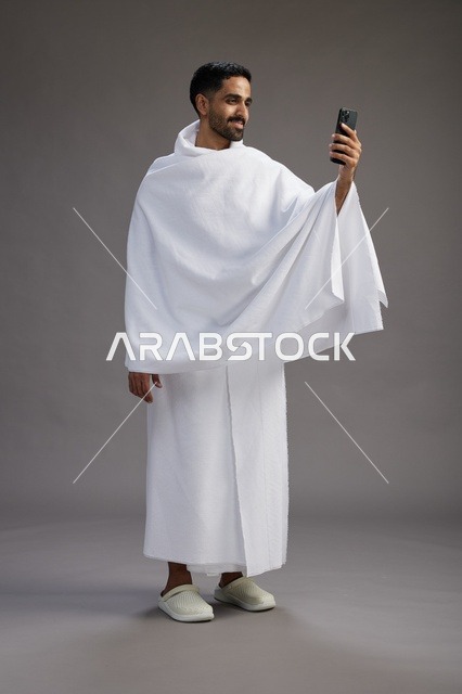 A portrait of a Saudi man in Ihram clothing smiling and taking a selfie, using a mobile phone, during Hajj and Umrah in Mecca, full body, gray background.