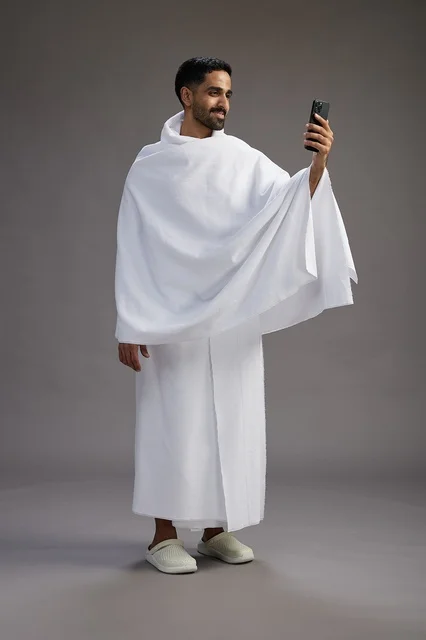 بورتريه لرجل سعودي بملابس الإحرام يبتسم ويلتقط صورة سيلفي, استخدام الهاتف المحمول, الحج والعمره في مكه, الجسم بالكامل, خلفيه رماديه