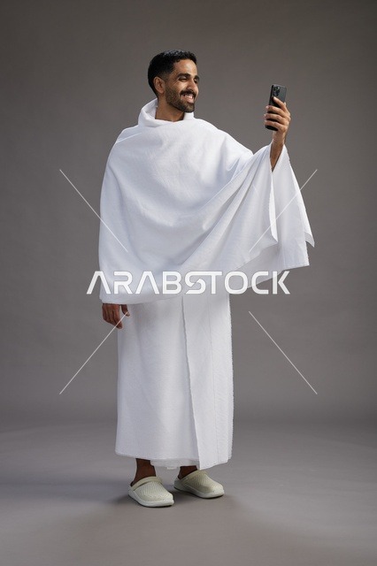 بورتريه لرجل سعودي بملابس الإحرام يبتسم ويلتقط صورة سيلفي, استخدام الهاتف المحمول, الحج والعمره في مكه, الجسم بالكامل, خلفيه رماديه