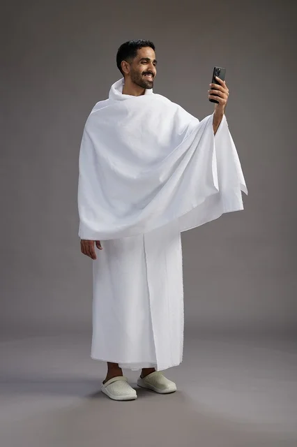 A portrait of a Saudi man in Ihram clothing smiling and taking a selfie, using a mobile phone, during Hajj and Umrah in Mecca, full body, gray background.