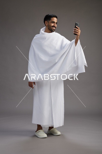 A portrait of a Saudi man in Ihram clothing smiling and taking a selfie, using a mobile phone, during Hajj and Umrah in Mecca, full body, gray background.