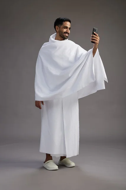 بورتريه لرجل سعودي بملابس الإحرام يبتسم ويلتقط صورة سيلفي, استخدام الهاتف المحمول, الحج والعمره في مكه, الجسم بالكامل, خلفيه رماديه