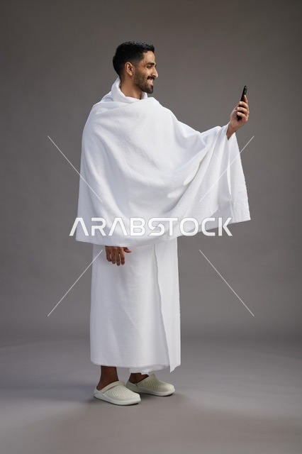 بورتريه لرجل سعودي بملابس الإحرام يبتسم ويلتقط صورة سيلفي, استخدام الهاتف المحمول, الحج والعمره في مكه, الجسم بالكامل, خلفيه رماديه