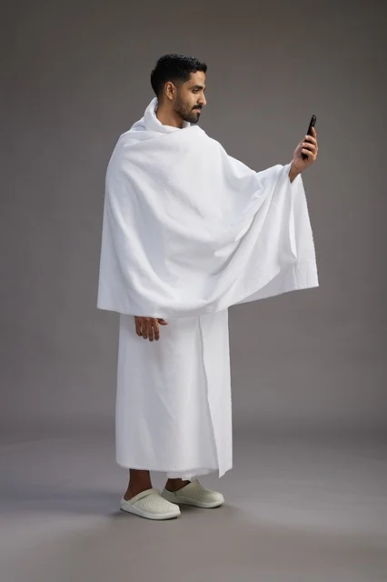 بورتريه لرجل سعودي بملابس الإحرام يبتسم ويلتقط صورة سيلفي, استخدام الهاتف المحمول, الحج والعمره في مكه, الجسم بالكامل, خلفيه رماديه