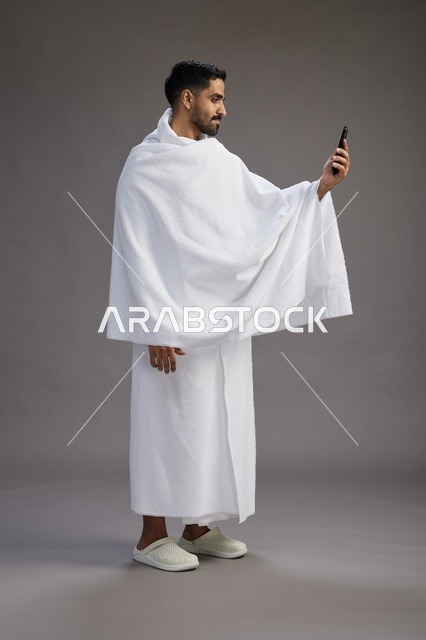 بورتريه لرجل سعودي بملابس الإحرام يبتسم ويلتقط صورة سيلفي, استخدام الهاتف المحمول, الحج والعمره في مكه, الجسم بالكامل, خلفيه رماديه