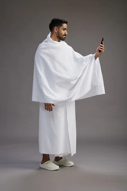 A portrait of a Saudi man in Ihram clothing smiling and taking a selfie, using a mobile phone, during Hajj and Umrah in Mecca, full body, gray background.